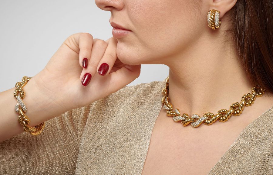 Una donna indossa una collana a catena in oro e diamanti, un bracciale abbinato e orecchini a cerchio di perle naturali, con la mano vicino al viso. Ha lo smalto rosso sulle unghie e indossa un top beige scintillante.