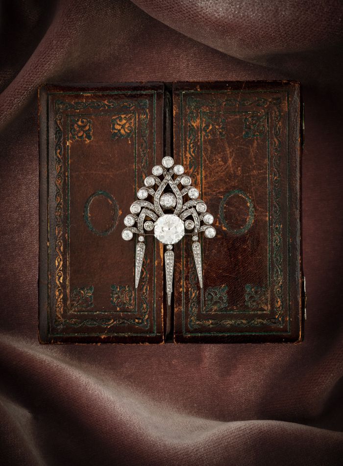 Una spilla vintage di diamanti e perle naturali è appoggiata su un vecchio libro rilegato in pelle con dettagli intricati. Lo sfondo è drappeggiato con un ricco tessuto marrone, creando un ambiente lussuoso ed elegante, perfetto per qualsiasi asta.
