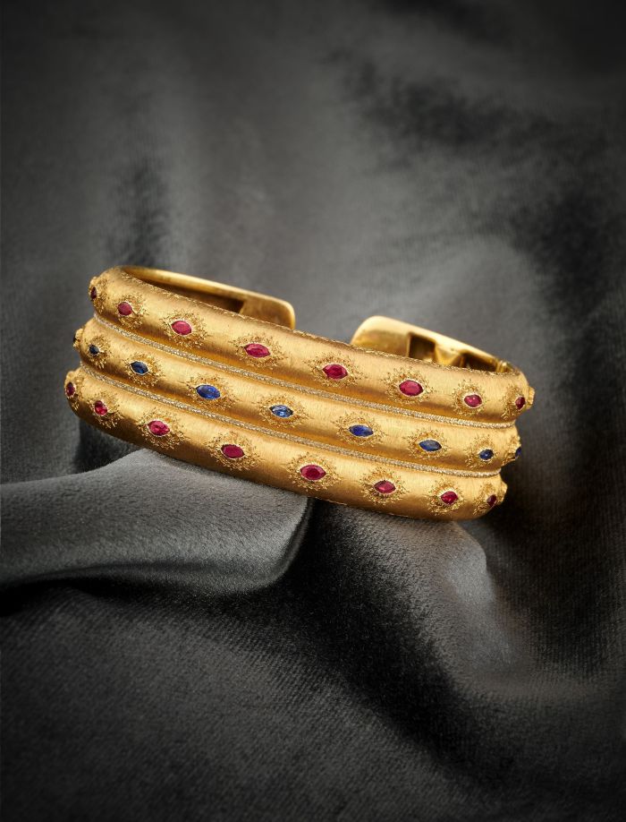 Un bracciale rigido in oro con tre fasce texturizzate, ciascuna tempestata di piccole pietre preziose rosse e blu e delicate perle naturali, su uno sfondo di tessuto grigio scuro drappeggiato.