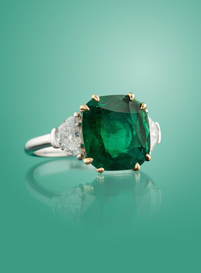 Un anello in argento con una grande pietra centrale di smeraldo verde taglio cuscino con punte in oro, affiancata da due diamanti trasparenti triangolari e delicate perle naturali, su uno sfondo verde sfumato.