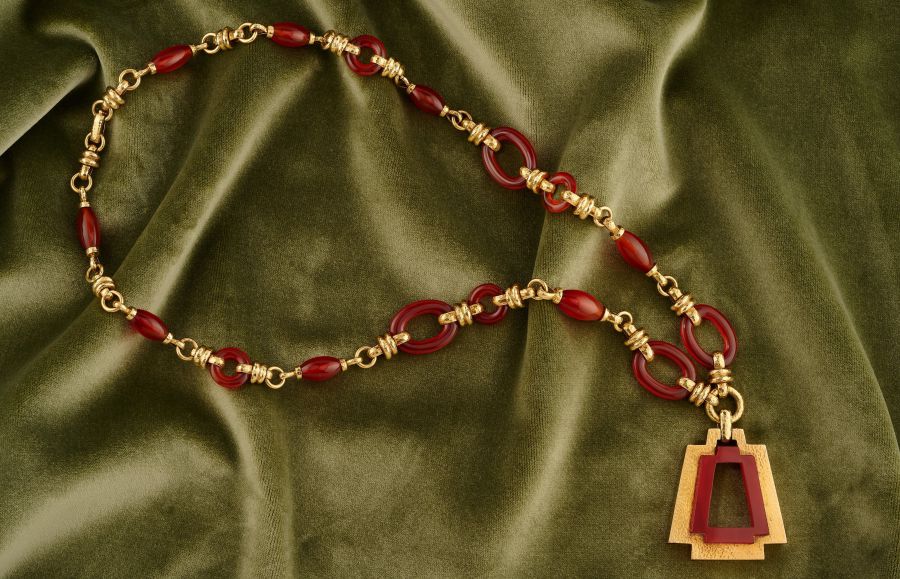 Collana con perline dorate e rosse, perle naturali e pendente geometrico, su uno sfondo in morbido velluto verde oliva.