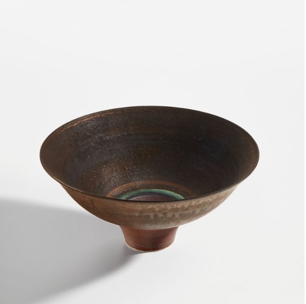 Una ciotola in ceramica ampia e poco profonda, con una superficie strutturata di colore marrone scuro e un piccolo piede circolare, mette in mostra le donne asta dal design esperto, elegantemente esposte su uno sfondo bianco.
