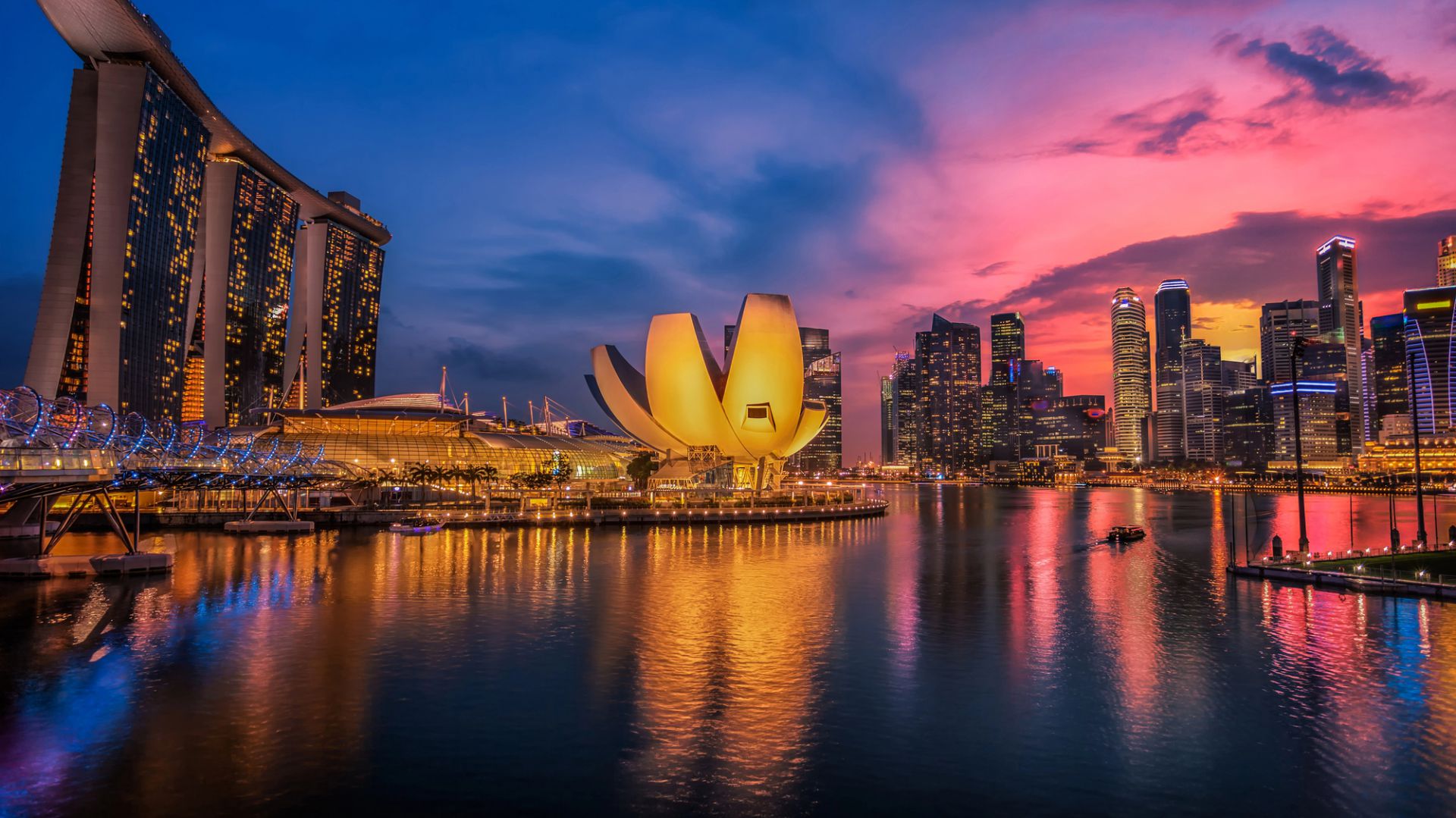 Lo skyline della città di Singapore al tramonto mostra edifici moderni e luminosi e simboli del lusso consumistico, tra cui l'iconico hotel Marina Bay Sands e l'ArtScience Museum, che si riflettono nella calma della baia sotto un vibrante cielo rosa e blu.