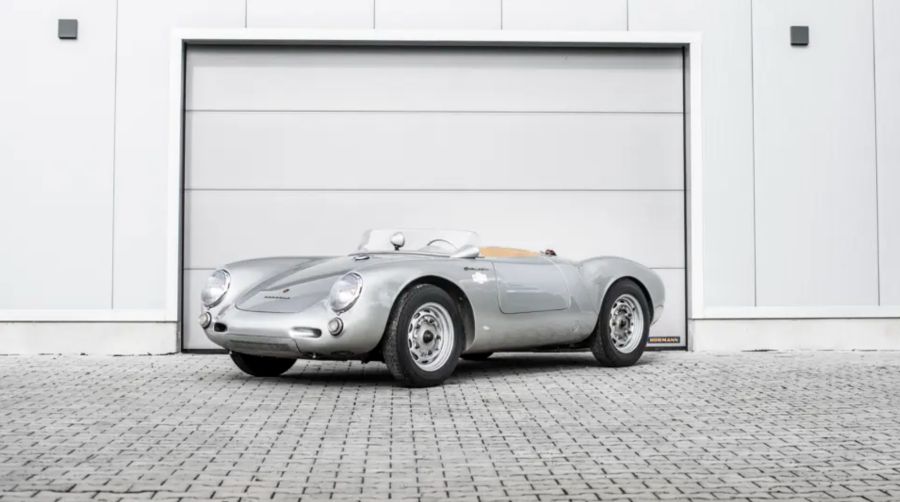 Una classica Porsche 550 Spyder decappottabile sportiva color argento, perfetta per la Mille Miglia 2025, è parcheggiata su un vialetto acciottolato di fronte a un moderno portone da garage grigio.