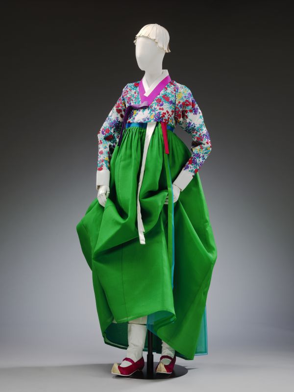 Un manichino vestito con il tradizionale hanbok coreano, ispirato all'ondata hallyu, presenta una gonna verde vivace, una giacca a motivi floreali con maniche lunghe, nastri bianchi e scarpe tradizionali rosse su uno sfondo grigio semplice.