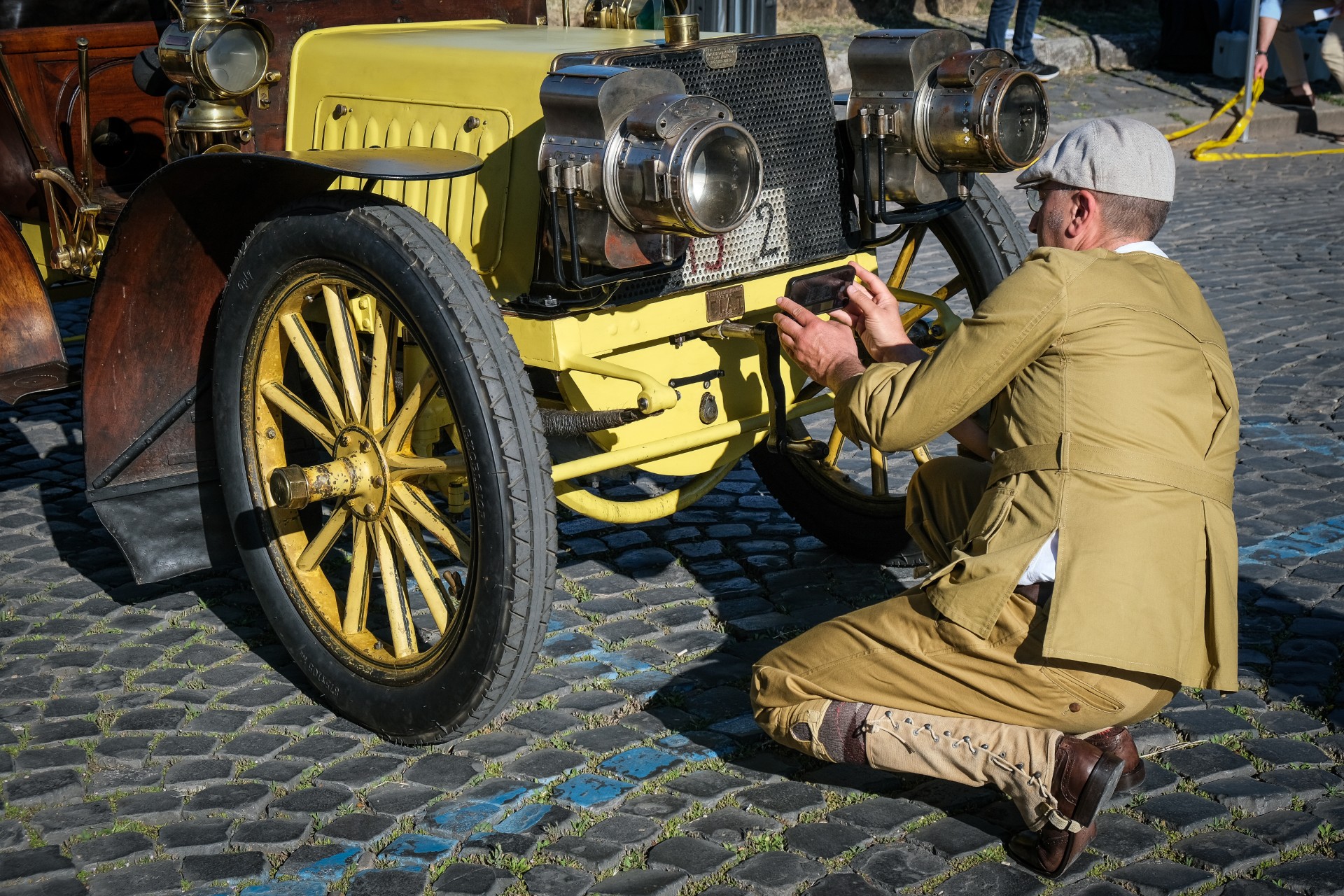Una persona in abiti d'epoca si inginocchia sul selciato, regolando la parte anteriore di un'auto d'epoca gialla con grandi fari e ruote a raggi di legno all'evento Auto storiche giubileo.