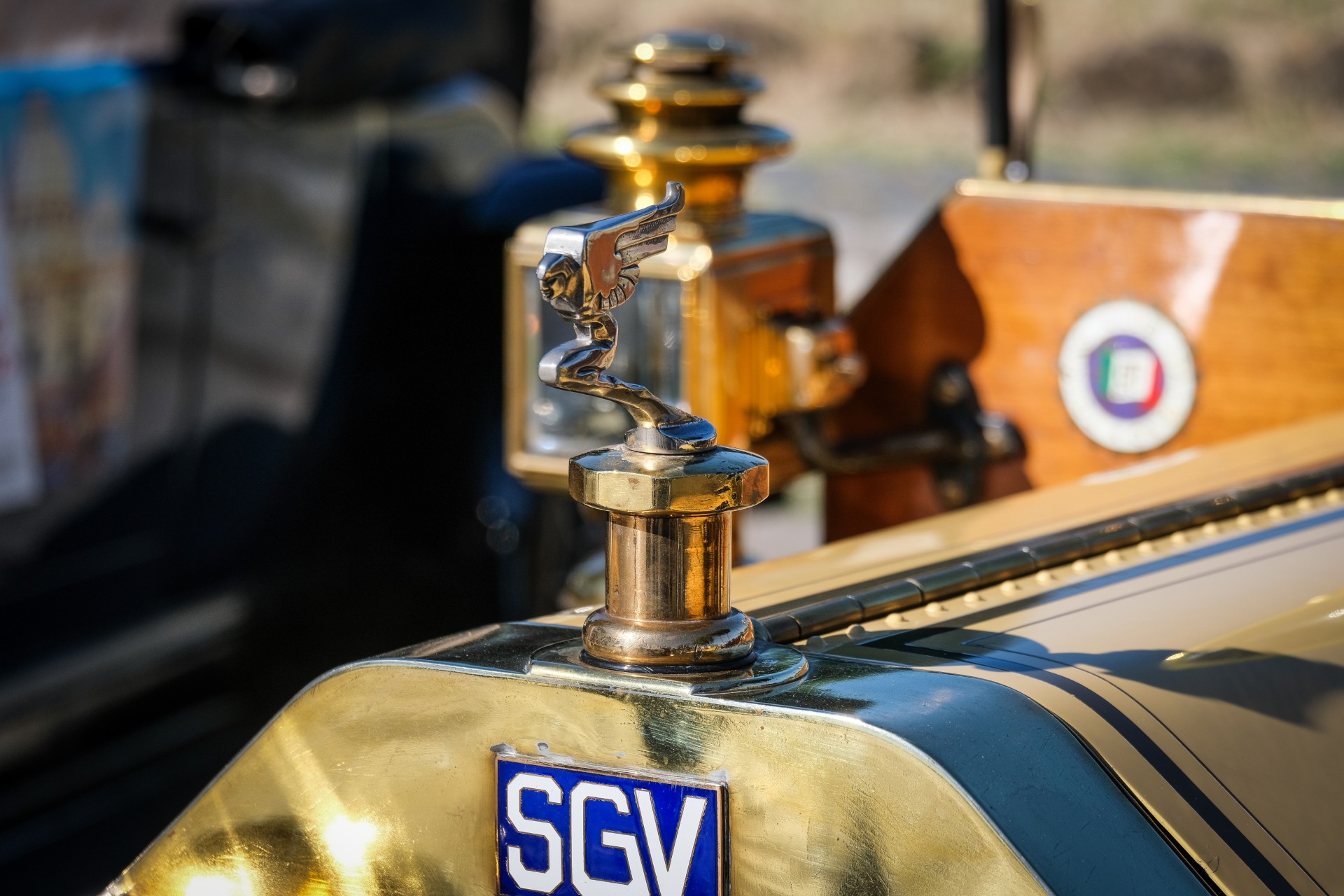 Primo piano dell'ornamento del cofano di un'auto d'epoca e dei dettagli in ottone, con targa blu "SGV", ripresi durante l'evento Auto storiche giubileo, con uno sfondo sfocato che mostra parti di un altro veicolo d'epoca.