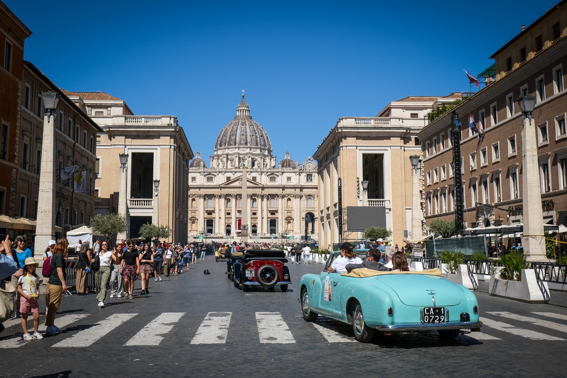 Auto storiche giubileo guida verso la Basilica di San Pietro nella Città del Vaticano, con folle di persone su entrambi i lati della strada e un cielo azzurro e limpido sopra di noi.