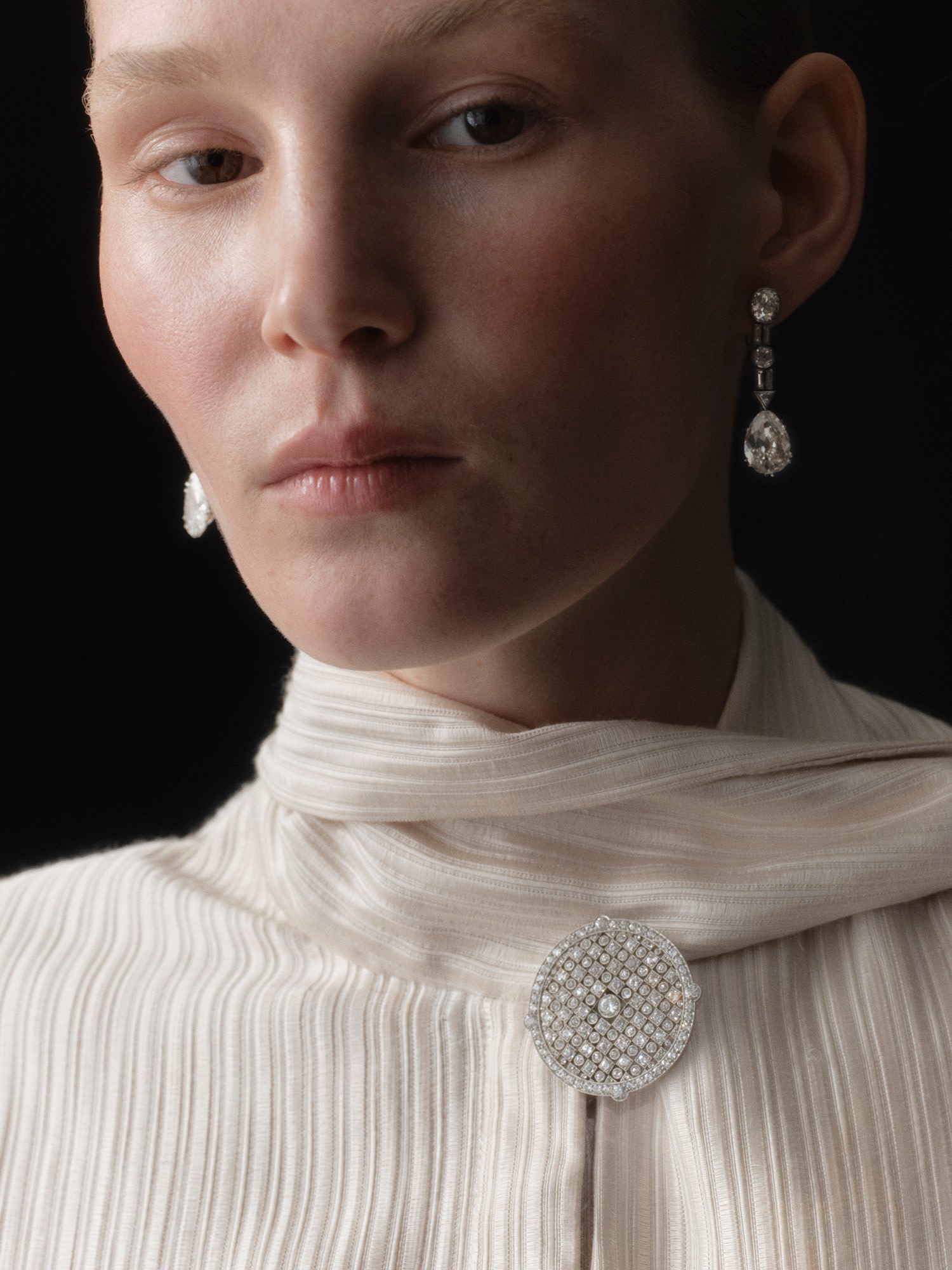 Una persona che indossa un indumento color crema testurizzato con una spilla di diamanti circolare e intricata, orecchini pendenti di diamanti abbinati e un elegante orologio Bulgari Serpente, posa su uno sfondo scuro.