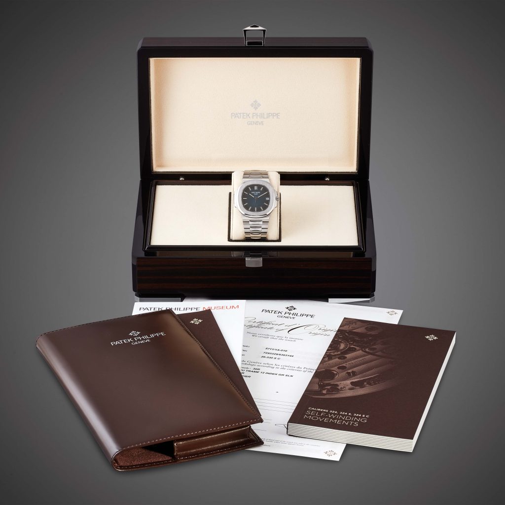Un lussuoso orologio Patek Philippe Nautilus asta in una scatola espositiva, con documenti marchiati, garanzia, libretto e una cartella in pelle marrone disposti ordinatamente sul davanti su uno sfondo neutro.
