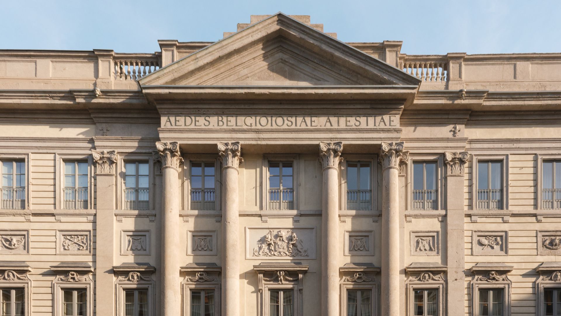 La facciata di un edificio neoclassico con alte colonne, intagli ornamentali, balconi e un frontone triangolare. Sopra le colonne è visibile l'iscrizione "AEDES BELGIOIOSIAE AESTIAE".