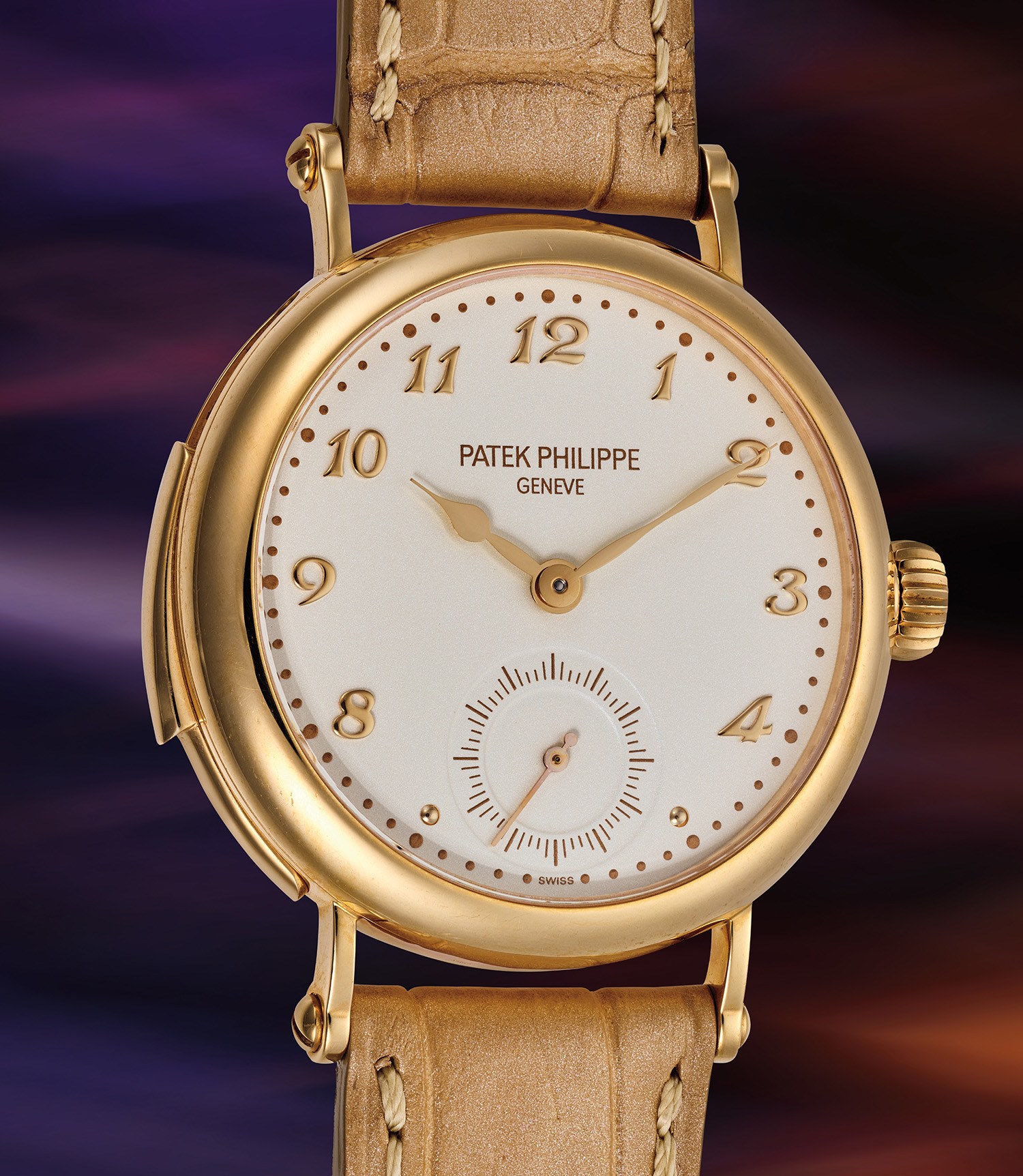 Un orologio da polso Patek Philippe in oro con cinturino in pelle marrone chiaro, numeri arabi e piccolo quadrante dei secondi è esposto in un espositore Phillips su uno sfondo sfocato viola scuro e rosa.