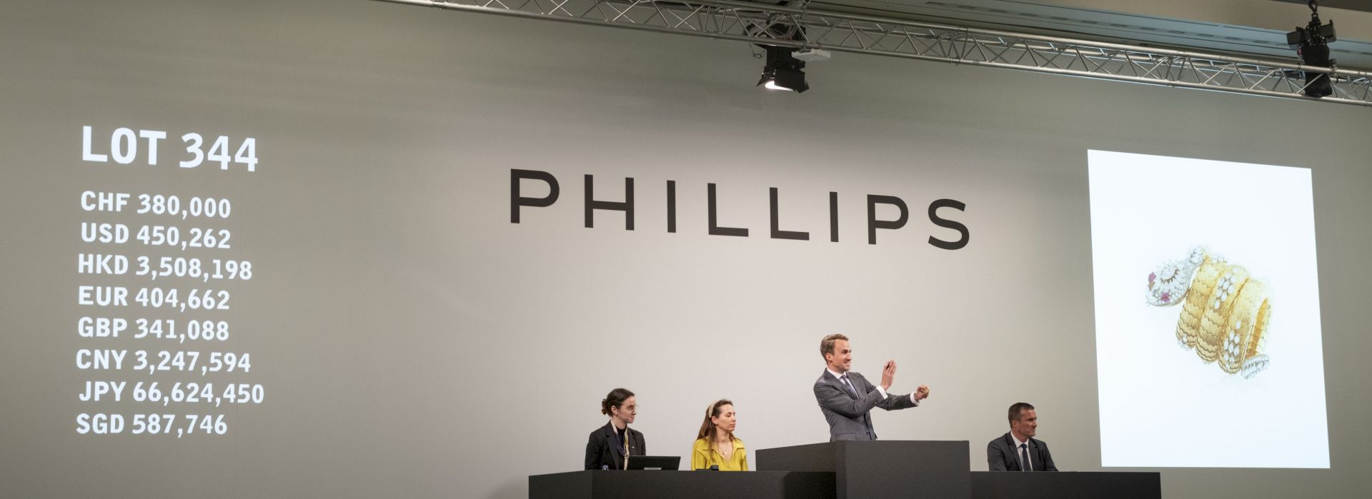 Un banditore della casa d'aste Phillips è in piedi su un podio e gesticola mentre guida le offerte per il lotto 344. Tre membri dello staff siedono accanto a lui e un grande schermo mostra un orologio Bulgari Serpente e i prezzi delle offerte in diverse valute.