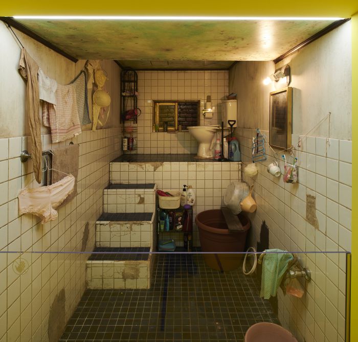 Un bagno piccolo e disordinato, con piastrelle bianche consumate, gradini che conducono alla toilette, biancheria appesa, materiale di pulizia sparso e accenni alla cultura hallyu nei poster alle pareti, il tutto sotto una luce fioca.