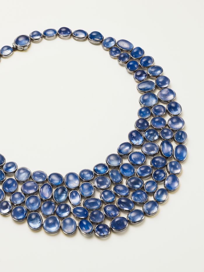 Primo piano di una collana con tre file di pietre preziose blu ovali incastonate in argento, disposte su uno sfondo bianco.