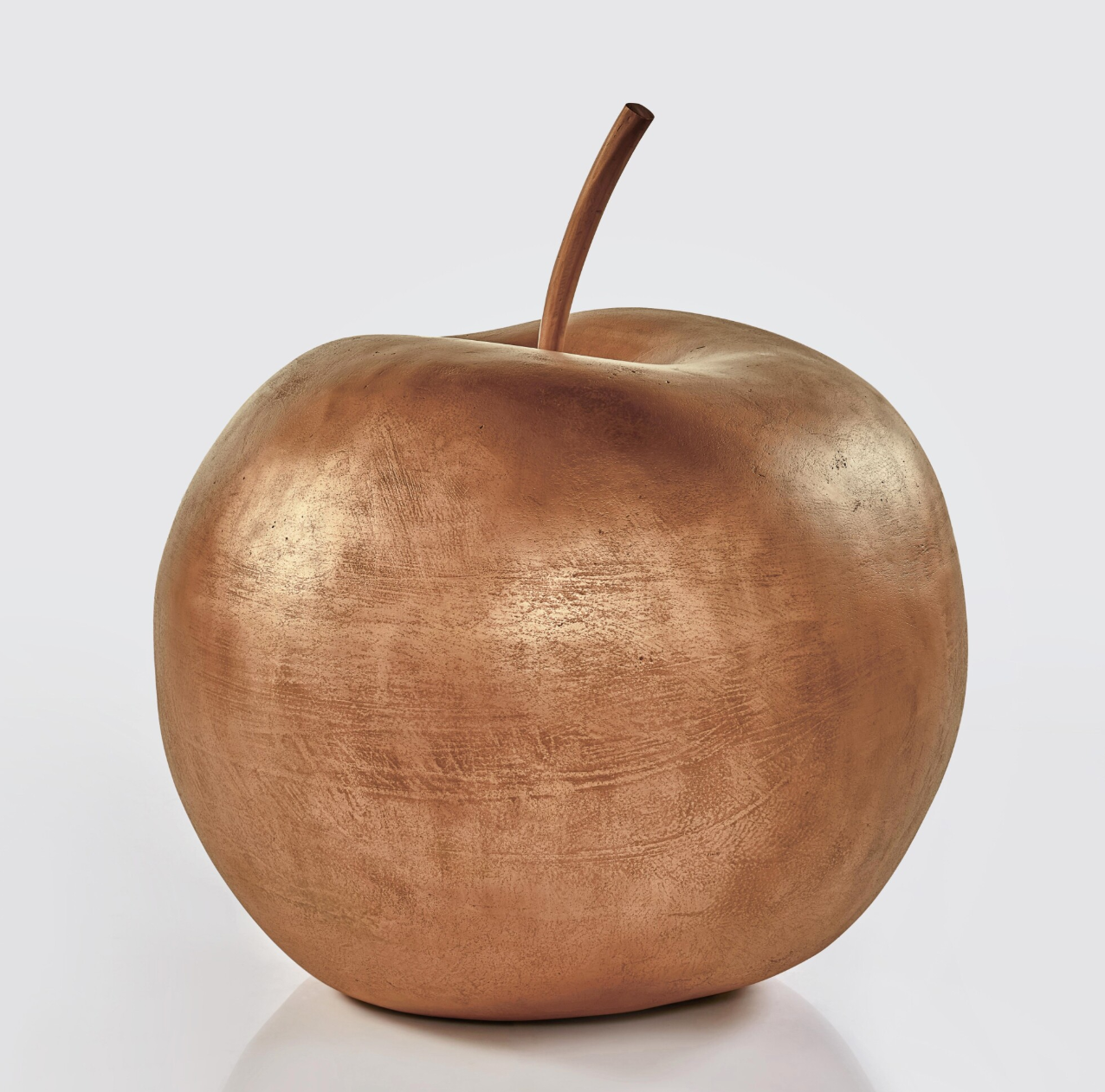 Una scultura metallica di grandi dimensioni raffigurante una mela con finitura liscia e lucida in bronzo e gambo ricurvo, esposta su un semplice sfondo bianco.