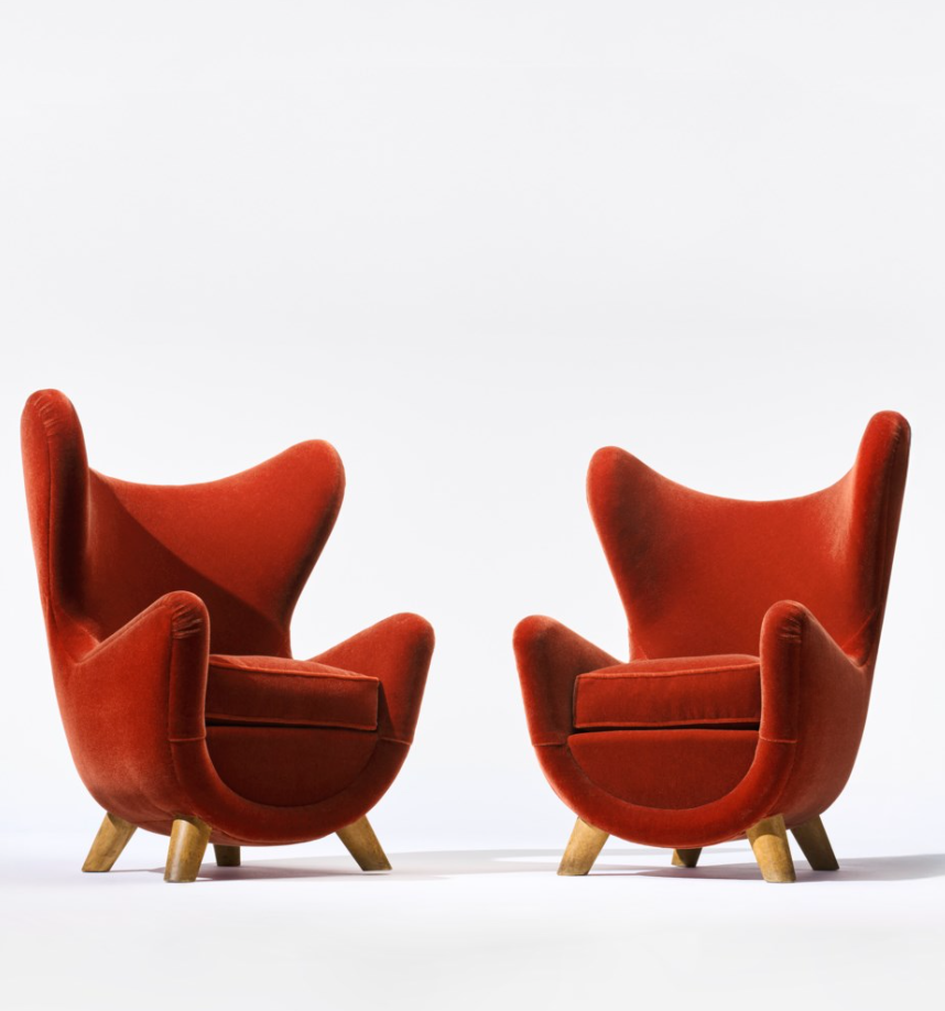 Due poltrone moderne della metà del secolo scorso con rivestimento in velluto rosso e schienali curvi e alati si trovano l'una di fronte all'altra su gambe di legno chiaro su uno sfondo bianco.