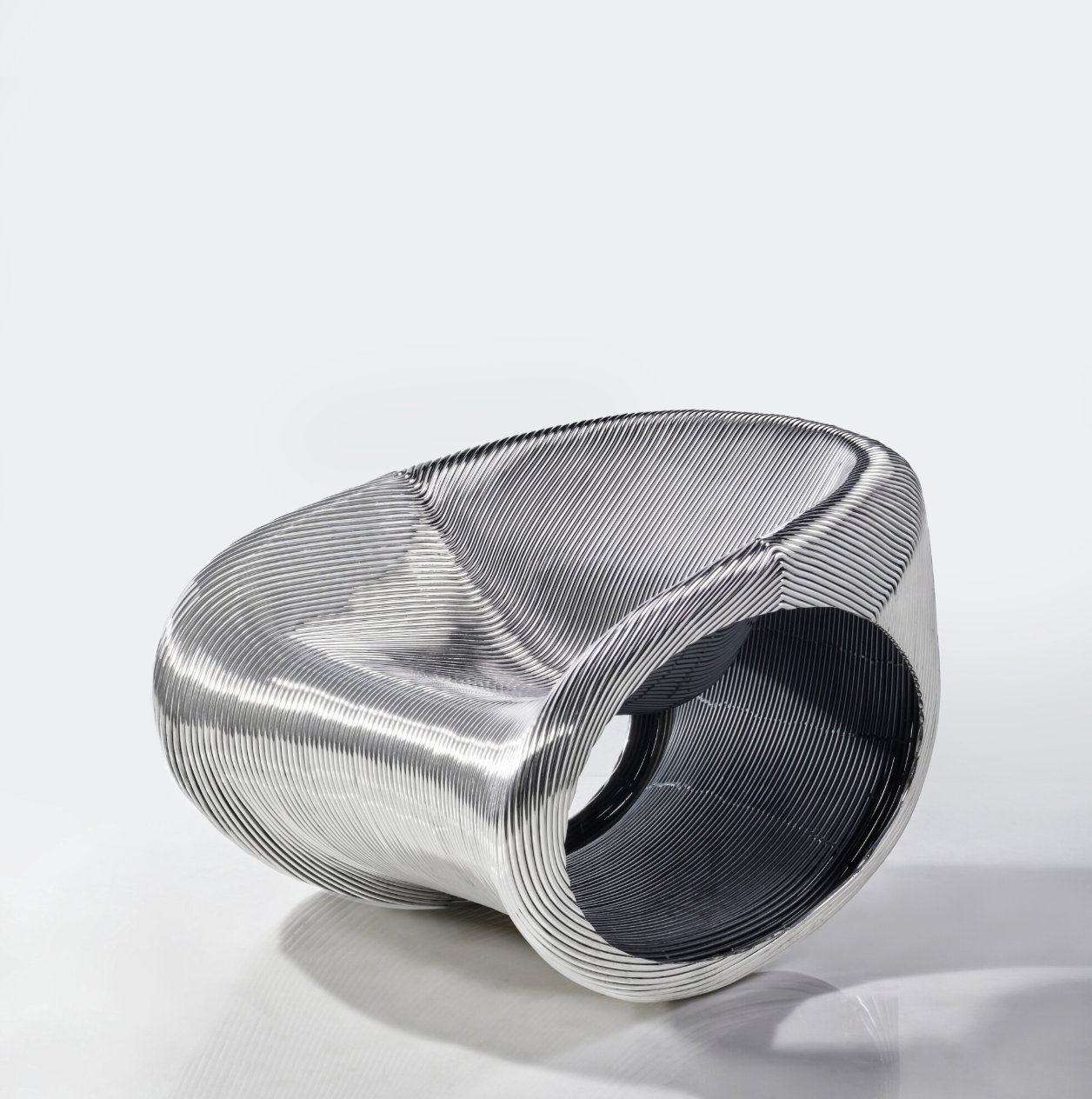 Una moderna poltrona metallica con base circolare e superficie curva e scanalata. Il design è caratterizzato da un fondo aperto e cavo e da un'elegante finitura argentata riflettente su uno sfondo bianco.