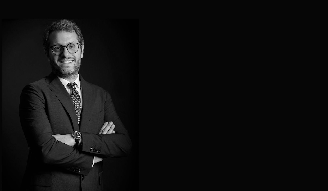 Foto in bianco e nero di un Simone Ragazzi, portfolio manager di Algebris Investments in giacca, cravatta e occhiali, in piedi con le braccia incrociate su uno sfondo scuro. Il lato destro dell'immagine è per lo più vuoto.