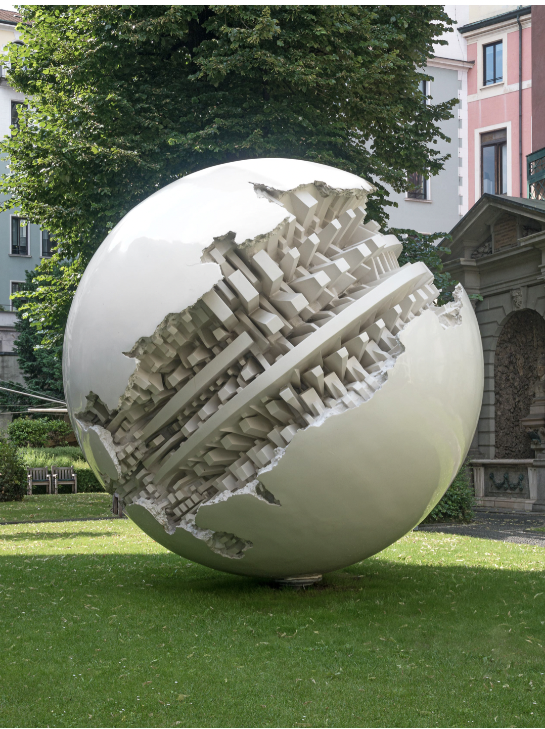 Una grande scultura a sfera metallica di Arnaldo Pomodoro, con ritagli geometrici frastagliati che rivelano intricati strati all'interno, esposta su un prato con alberi ed edifici sullo sfondo.