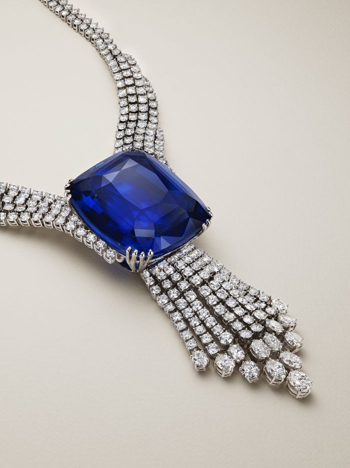 Una lussuosa collana con al centro una grande pietra preziosa blu intenso, circondata da più file di diamanti scintillanti incastonati in argento, su uno sfondo beige chiaro.