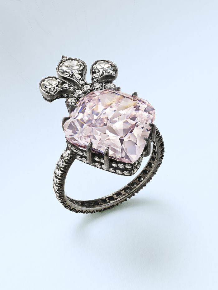 Un anello d'argento con una grande pietra preziosa rosa pallido al centro, impreziosita da pietre trasparenti più piccole disposte a formare un fiocco decorativo sopra la gemma principale, fotografato su uno sfondo chiaro.