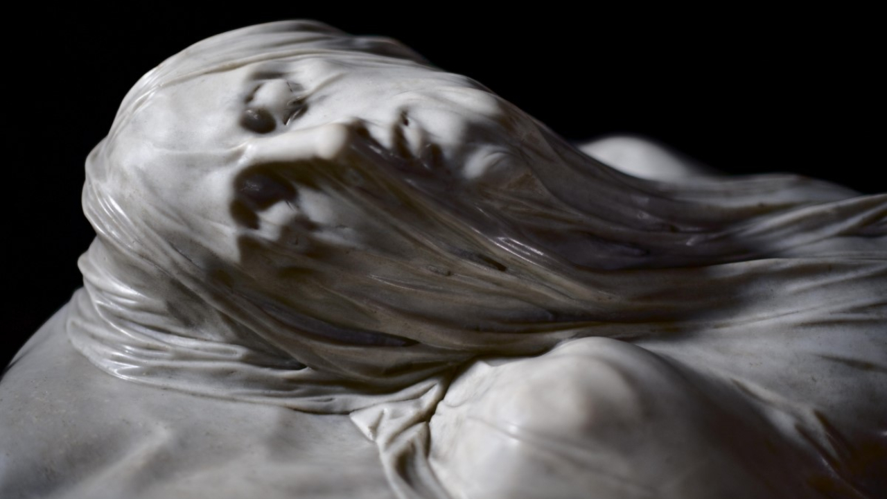Primo piano di una scultura in marmo, ispirata all'arte napoletana, che raffigura una figura reclinata con un velo trasparente drappeggiato sul viso e sul busto, intricatamente scolpito per rivelare i dettagli del viso sotto il tessuto.