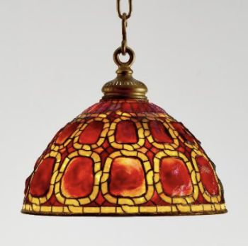 Una lampada a sospensione in vetro colorato con paralume a forma di cupola con motivi geometrici rossi e gialli, sospesa da una catena in ottone, mette in mostra un elegante design femminile su uno sfondo semplice.