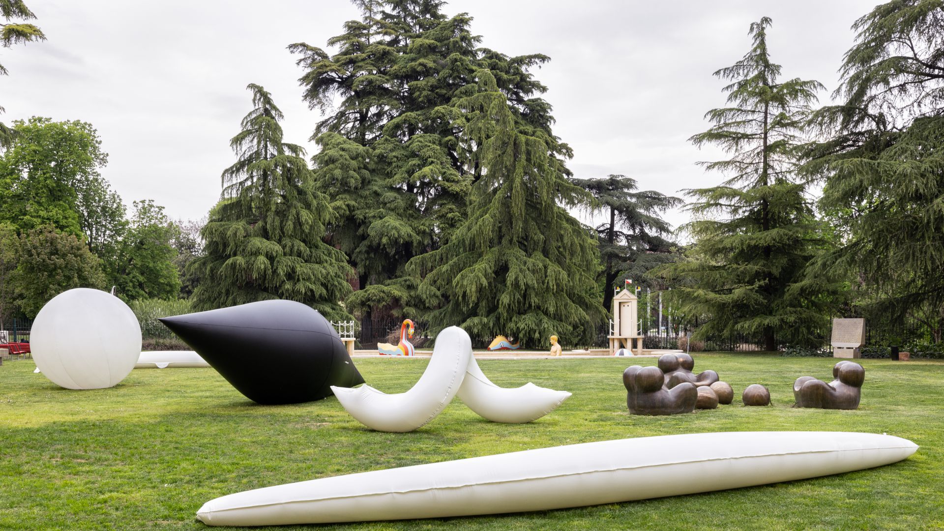 Grandi sculture astratte di varie forme, tra cui una goccia nera, una sfera bianca, forme curve e forme organiche marroni, sono esposte su un prato circondato da alti alberi verdi.