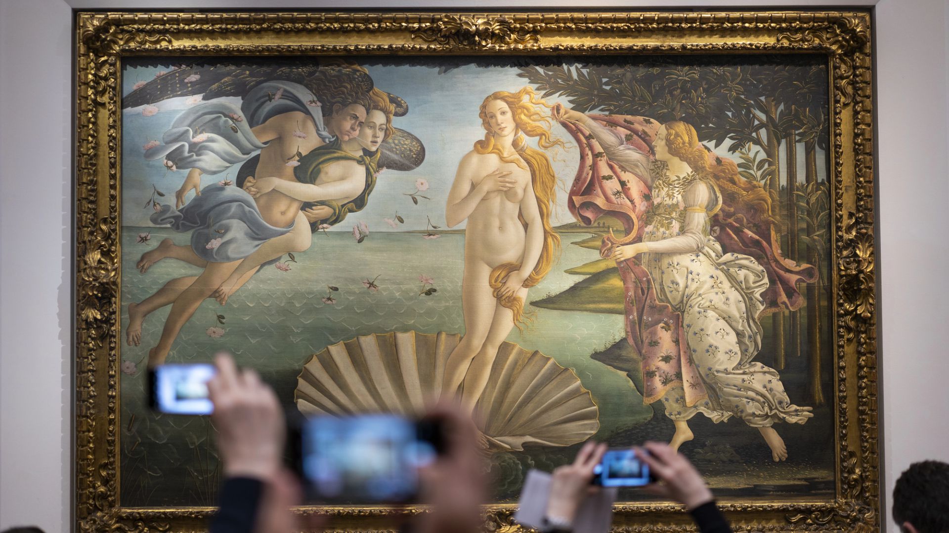 I turisti si scattano un selfie davanti alla "Nascita di Venere" di Sandro Botticelli agli Uffizi, immortalando il famoso dipinto rinascimentale di Venere che emerge da una conchiglia tra figure mitologiche in una cornice dorata.