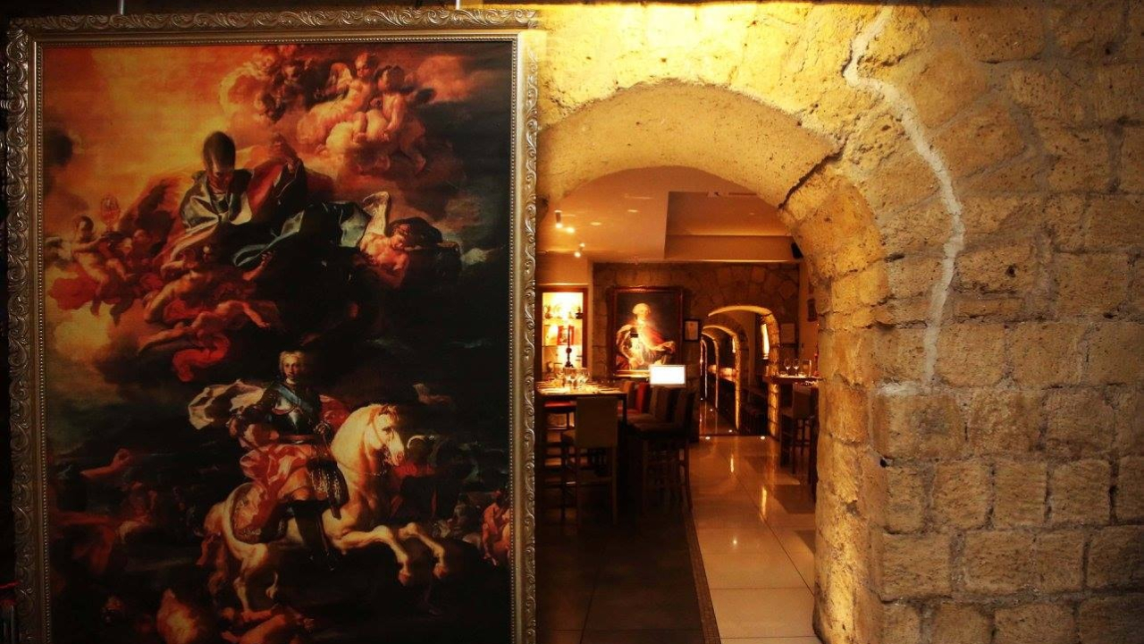 Un grande quadro classico incorniciato è appeso a una parete di pietra vicino a una porta ad arco. Attraverso l'arco, sullo sfondo si intravede un ristorante Napoli illuminato con tavoli e sedie in legno.