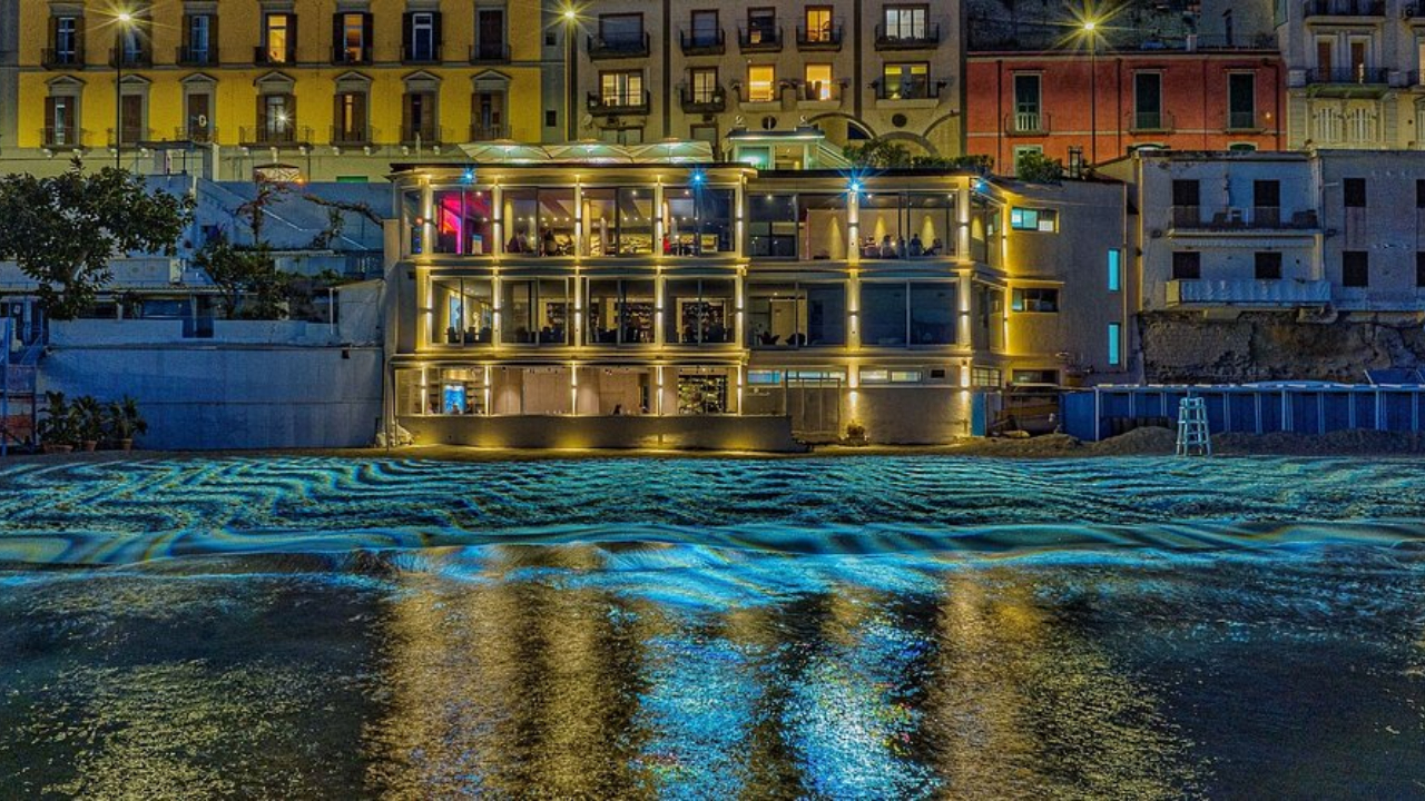 Un edificio moderno e ben illuminato con grandi finestre si trova sul lungomare di Napoli di notte, riflettendo luci dorate e blu sull'acqua increspata; alle sue spalle sorgono edifici colorati della città.