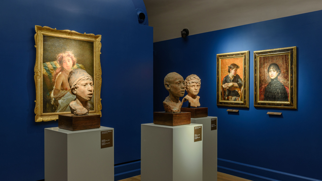 In una sala della galleria d'arte di Napoli, con pareti di un blu intenso, sono esposti tre busti scolpiti su piedistalli e tre ritratti incorniciati, ciascuno illuminato da faretti.