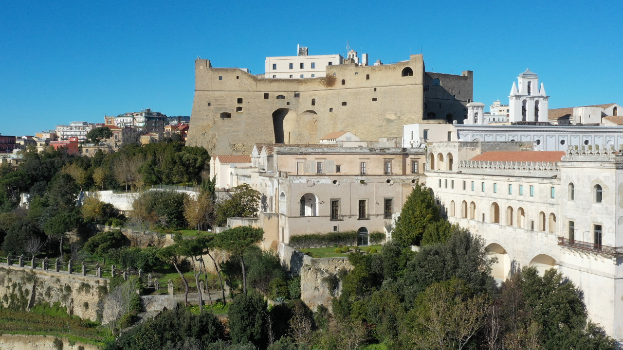 Una grande fortezza storica con mura di pietra beige si trova in cima a una collina di Napoli, circondata da alberi ed edifici bianchi, sotto un cielo azzurro e limpido.
