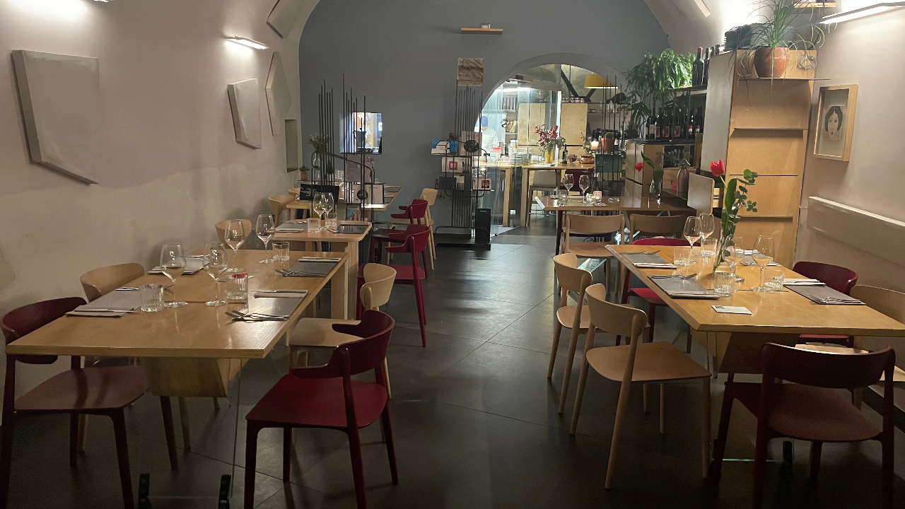 Un ristorante accogliente e moderno ispirato a Napoli, con tavoli in legno, sedie rosse e in legno apparecchiate per la cena con bicchieri e tovaglioli. Luci soffuse e piante creano un'atmosfera calda; la cucina è visibile sullo sfondo.