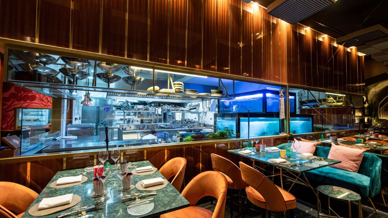 Un ristorante moderno ispirato a Napoli, con tavoli eleganti, sedie arancioni e verde acqua, cuscini rosa e un'ampia vetrata che rivela una luminosa e vivace cucina a vista con attrezzature in acciaio inox e acquari illuminati di blu.