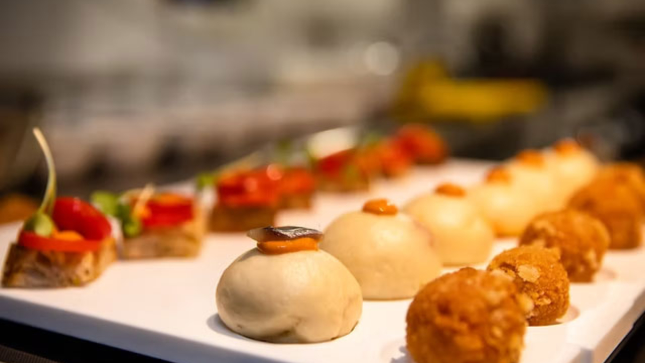 Primo piano di un vassoio bianco che espone vari stuzzichini, tra cui tartine salate di ispirazione napoletana, panini rotondi al vapore con guarnizioni e palline fritte dorate, su uno sfondo sfocato.