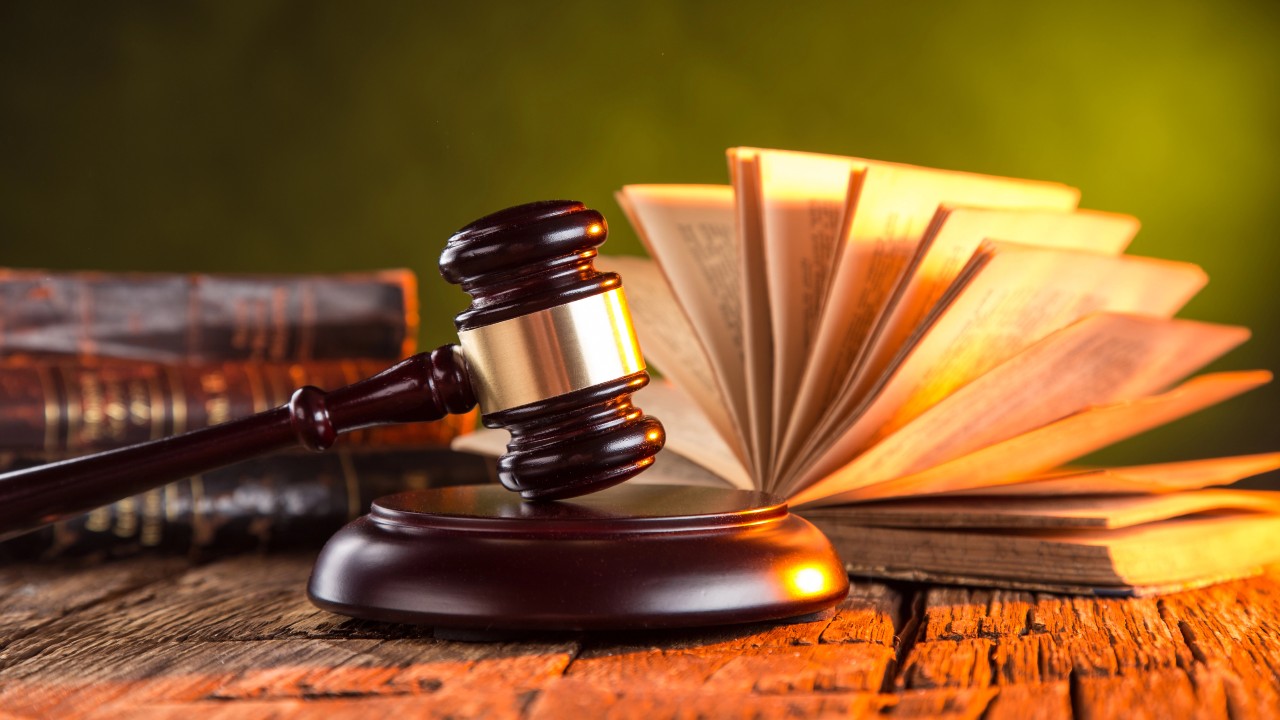 Un martelletto di legno poggia su un blocco sonoro davanti a un libro aperto e a libri giuridici impilati, che simboleggiano la lotta contro l'abuso del diritto.