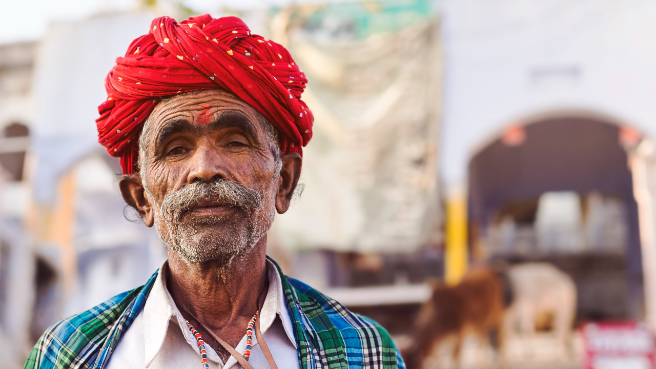 Un uomo anziano con baffi e barba che indossa un turbante rosso acceso e abiti tradizionali si trova all'aperto, con edifici sfocati e una mucca sullo sfondo.