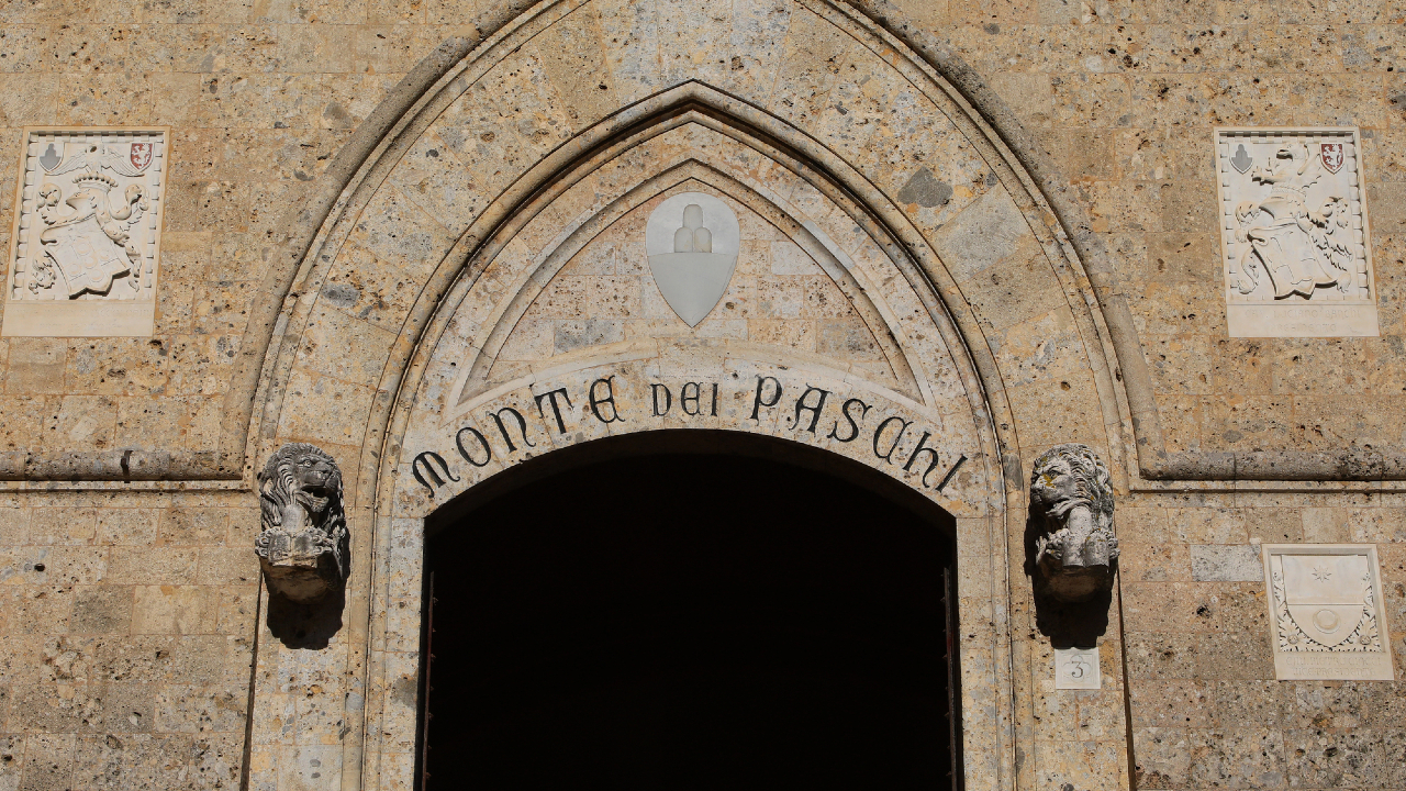 Un arco in pietra con due sculture a testa di leone e la scritta "Monte dei Paschi" scolpita sopra l'ingresso, inserito in un antico edificio in pietra con rilievi decorativi su entrambi i lati.