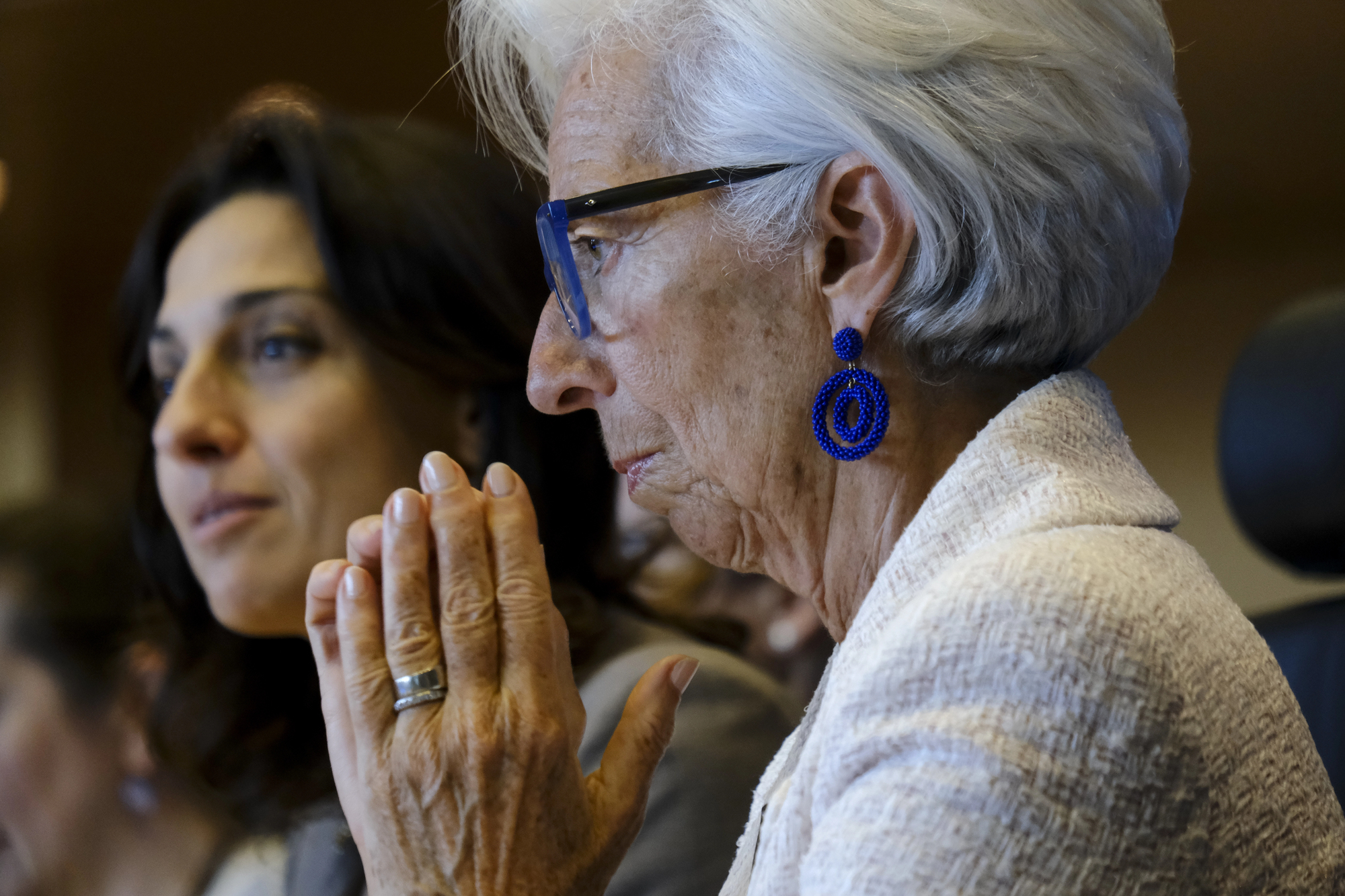 Due donne siedono una accanto all'altra, una con i capelli grigi, orecchini blu e occhiali, e tiene le mani giunte davanti al viso in un gesto pensieroso; l'altra donna appare leggermente sfocata sullo sfondo.