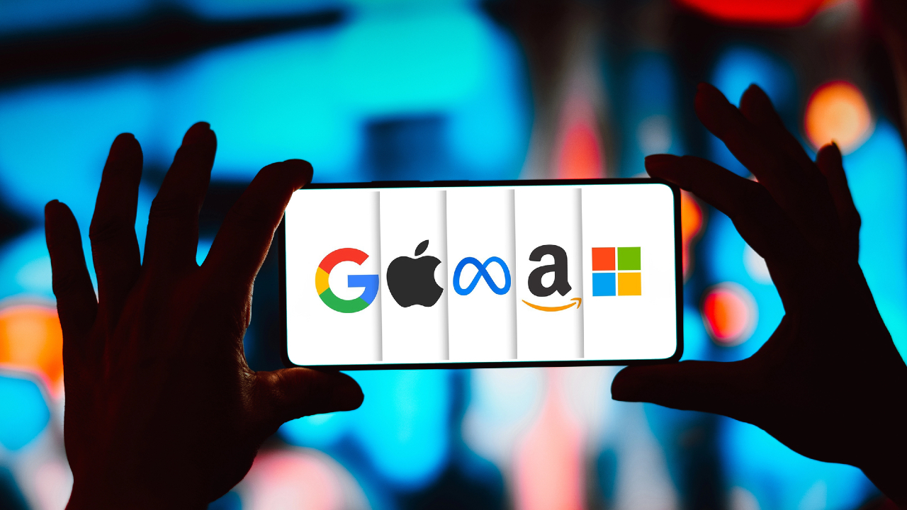 Una persona tiene in mano uno smartphone con i loghi dei grandi colossi tecnologici - Google, Apple, Meta, Amazon e Microsoft - su uno sfondo colorato e sfocato.