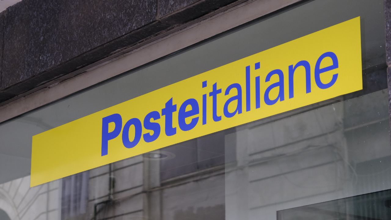 Un'insegna gialla con testo blu recita "Posteitaliane", indicando un ufficio postale italiano, esposto su una vetrata con riflessi di edifici vicini.