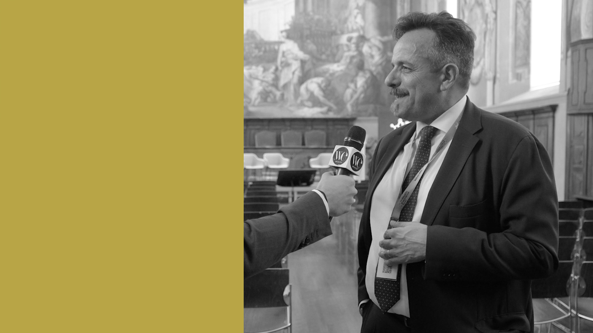 Berardo Staglianò in giacca e cravatta viene intervistato con un microfono in una stanza ornata e classica. Il lato sinistro dell'immagine è coperto da un blocco giallo senape.