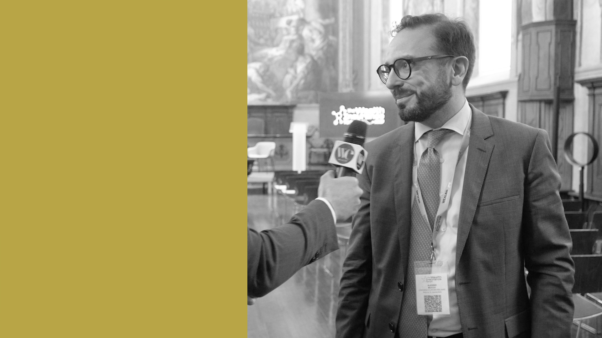 Alessio Bucco in abito e occhiali viene intervistato con un microfono in una stanza ornata; indossa un cordino e un cartellino con il nome. Il lato sinistro dell'immagine è coperto da un blocco di colore oro massiccio.