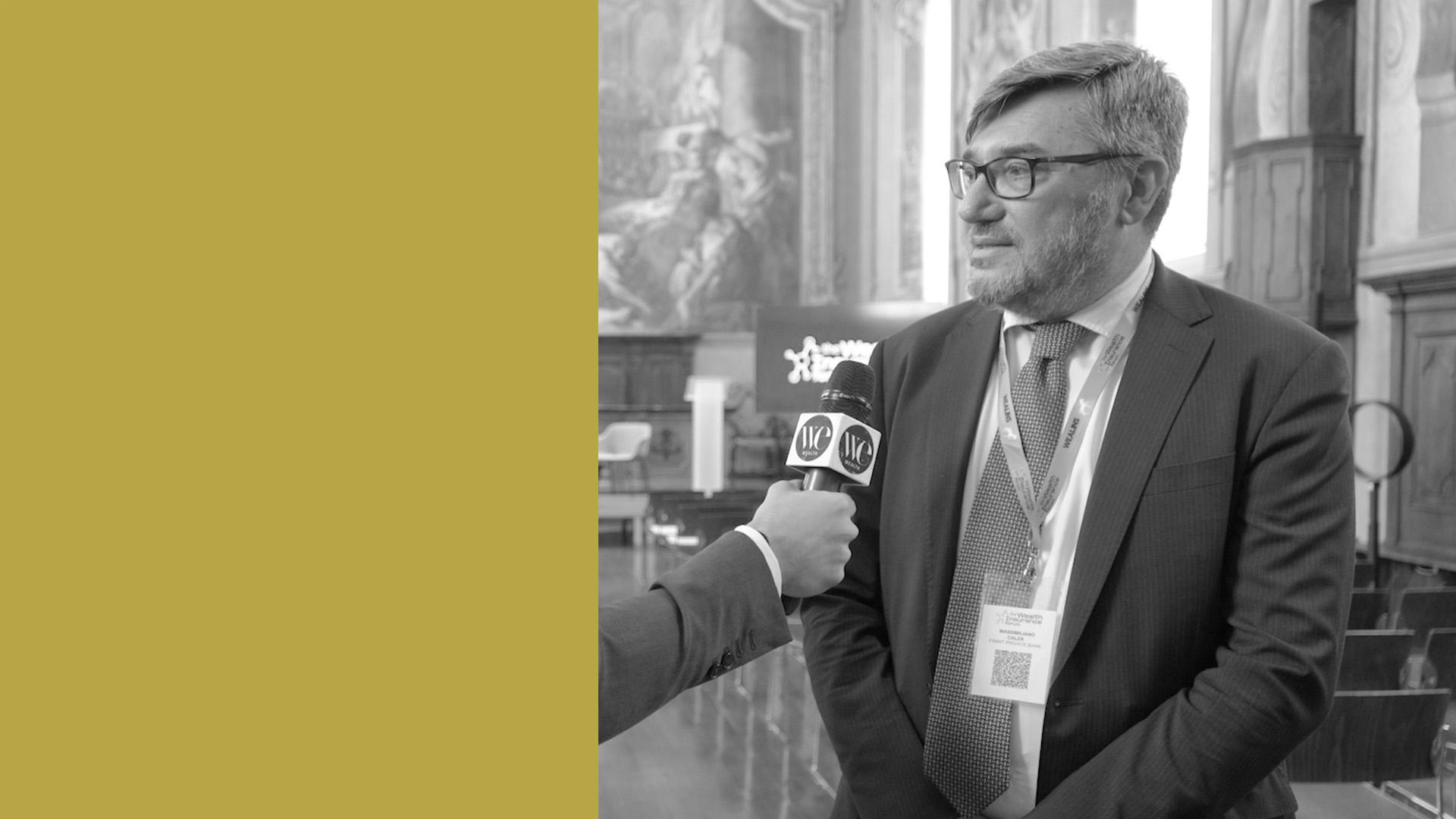 Un uomo in giacca e cravatta viene intervistato con un microfono in una grande sala dalle pareti decorate. Sul lato sinistro dell'immagine è presente un grande blocco giallo senape. La scena è in bianco e nero.