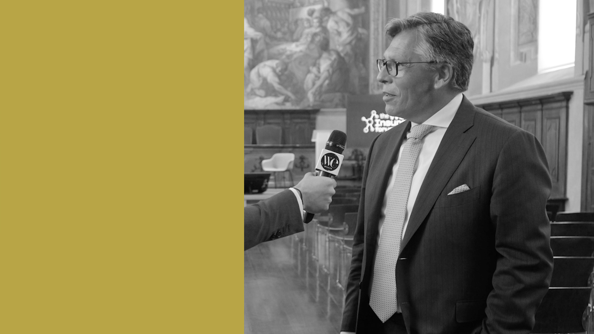 Luc Rasschaert in abito e occhiali viene intervistato in una stanza formale con opere d'arte decorate alle pareti. Il lato sinistro dell'immagine presenta un blocco di colore giallo senape.