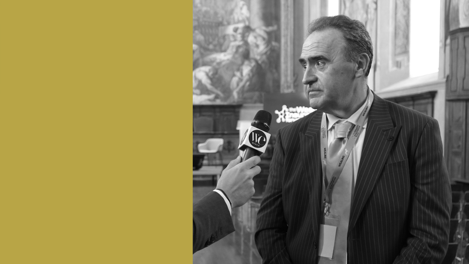 Angelo Taffurelli in giacca e cravatta viene intervistato con un microfono davanti a sé. Lo sfondo mostra una stanza decorata con opere d'arte. Il lato sinistro dell'immagine è coperto da un blocco giallo senape.