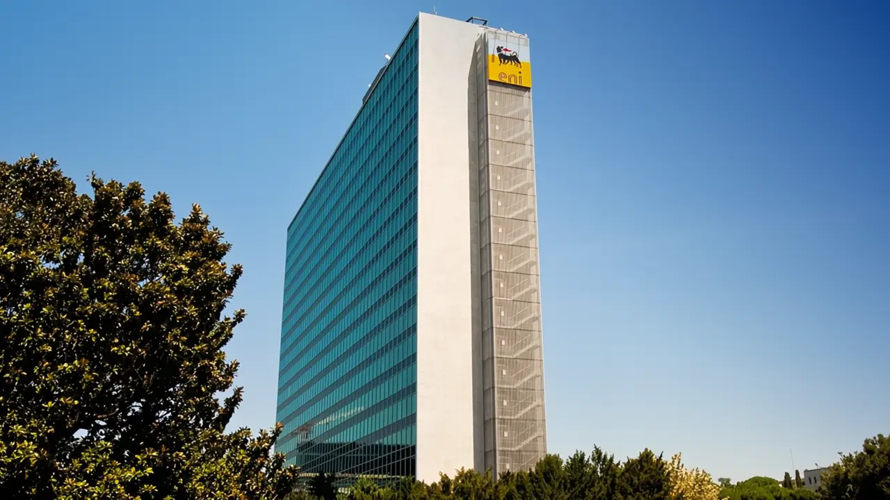 Un alto e moderno edificio per uffici in vetro con il logo dell'azienda Eni in cima si staglia contro un cielo azzurro e circondato da alberi verdi alla base.