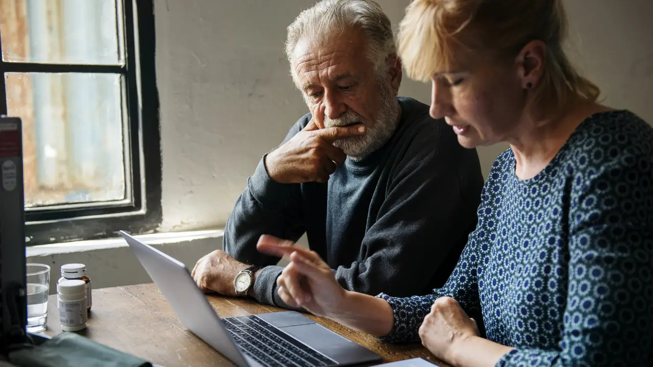 Un uomo e una donna anziani siedono a un tavolo di legno e guardano un computer portatile. La donna indica lo schermo mentre l'uomo ascolta pensieroso. Sul tavolo vicino a loro sono visibili dei flaconi di medicinali.